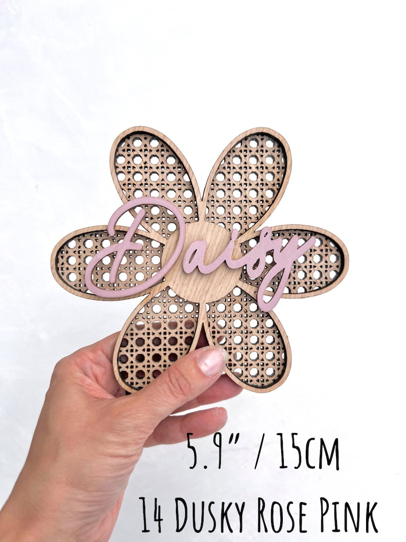 K&ouml;nnte beinhalten: Ein h&ouml;lzernes Schild in Form einer Blume mit einem gewebten Muster und dem Namen "Daisy" in Pink darauf. Das Schild hat einen Durchmesser von 15 cm und ist in einem matten Rosaton lackiert.