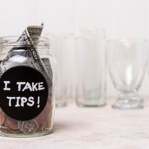 Op de afbeelding: Een glazen pot met een zwart label waarop "I take tips!" staat. De pot is gevuld met Amerikaanse dollarbiljetten en munten. Er staan vier lege glazen achter de pot.