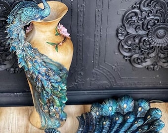 Puede incluir: Un jarrón decorativo con forma de pavo real con plumas azules y verdes. El jarrón está hecho de cerámica y tiene una base de color dorado. El jarrón está sobre una superficie de madera.