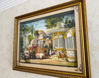 Op de afbeelding: Een ingelijst kunstwerk dat een gedetailleerd tafereel van een oude stad met gebouwen, bomen en mensen afbeeldt. Het kunstwerk is in een gouden lijst met sierlijke details.