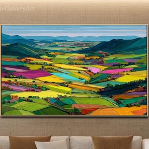 Pode incluir: Uma pintura de paisagem emoldurada com uma representação vibrante e colorida de colinas e campos. A obra de arte apresenta uma paleta de verdes, amarelos, rosas e azuis, criando um efeito de retalhos. A pintura é exibida acima de um sofá branco com almofadas marrons e brancas.
