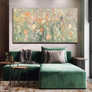 Op de afbeelding: Een abstract bloemenschilderij met een gouden lijst. Het kunstwerk toont een veld met bloemen in tinten oranje, roze en geel tegen een groen-witte achtergrond. Het schilderij hangt boven een groene fluwelen hoekbank met sierkussens.
