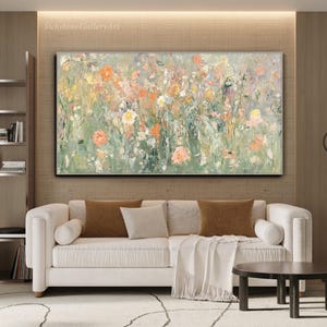 Op de afbeelding: Abstract bloemenschilderij met een zilveren lijst. Het kunstwerk toont een veld met bloemen in tinten oranje, geel, roze en wit tegen een groene en grijze achtergrond. Het schilderij wordt boven een witte bank tentoongesteld.