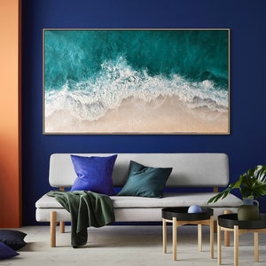 May include: Large rectangular framed artwork depicting an aerial view of a beach. The artwork features turquoise ocean waves crashing onto a sandy shore. The frame is a light brown color. The artwork is displayed above a gray sofa with blue and green pillows.