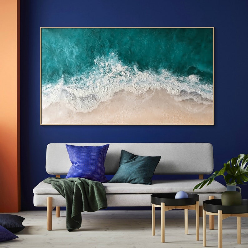 May include: Large rectangular framed artwork depicting an aerial view of a beach. The artwork features turquoise ocean waves crashing onto a sandy shore. The frame is a light brown color. The artwork is displayed above a gray sofa with blue and green pillows.