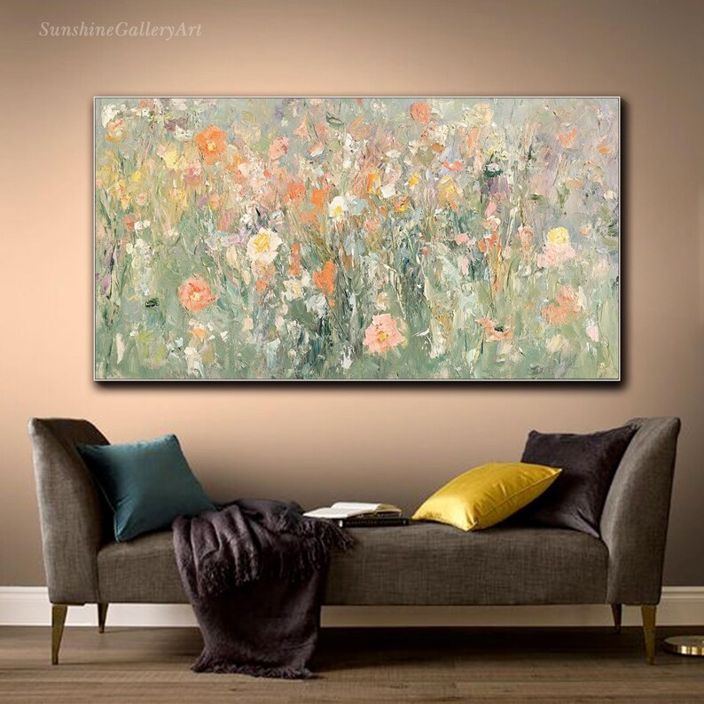 Op de afbeelding: Een abstract bloemenschilderij met zachte groen-, oranje- en roze tinten. Het kunstwerk heeft een textuur, impressionistische stijl, die een bloemenveld uitbeeldt. Het schilderij wordt tentoongesteld boven een grijze bank met kleurrijke kussens.