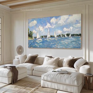 Op de afbeelding: Een schilderij van een zeegezicht met zeilboten op een blauwe watermassa. De lucht is blauw met witte wolken. Het schilderij is ingelijst en hangt aan een witte muur boven een witte bank.