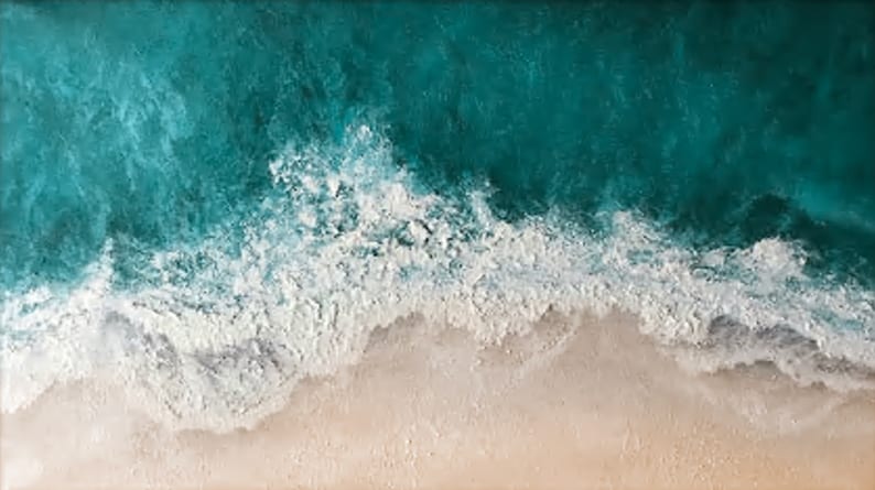 May include: An aerial view of a beach scene. The top portion of the image shows turquoise ocean water with white foamy waves crashing onto a sandy beach. The sand is a light tan color.