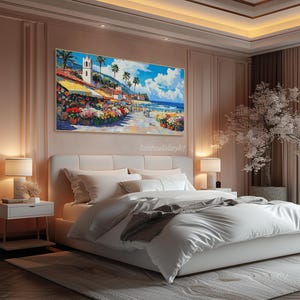 May include: A painting of a colorful seaside town with a white building and palm trees. The painting is hanging on a wall above a white bed with white bedding and a gray throw blanket. The room is decorated with a pink wall and a white nightstand.