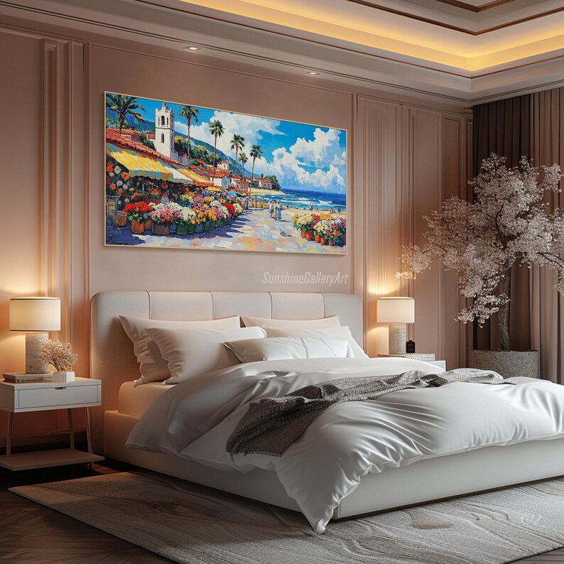 May include: A painting of a colorful seaside town with a white building and palm trees. The painting is hanging on a wall above a white bed with white bedding and a gray throw blanket. The room is decorated with a pink wall and a white nightstand.
