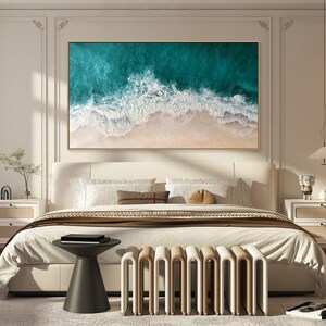May include: A framed artwork depicting an aerial view of ocean waves crashing on a sandy beach. The painting features shades of teal and white for the water, and beige for the sand. The artwork is displayed above a bed in a bedroom setting.