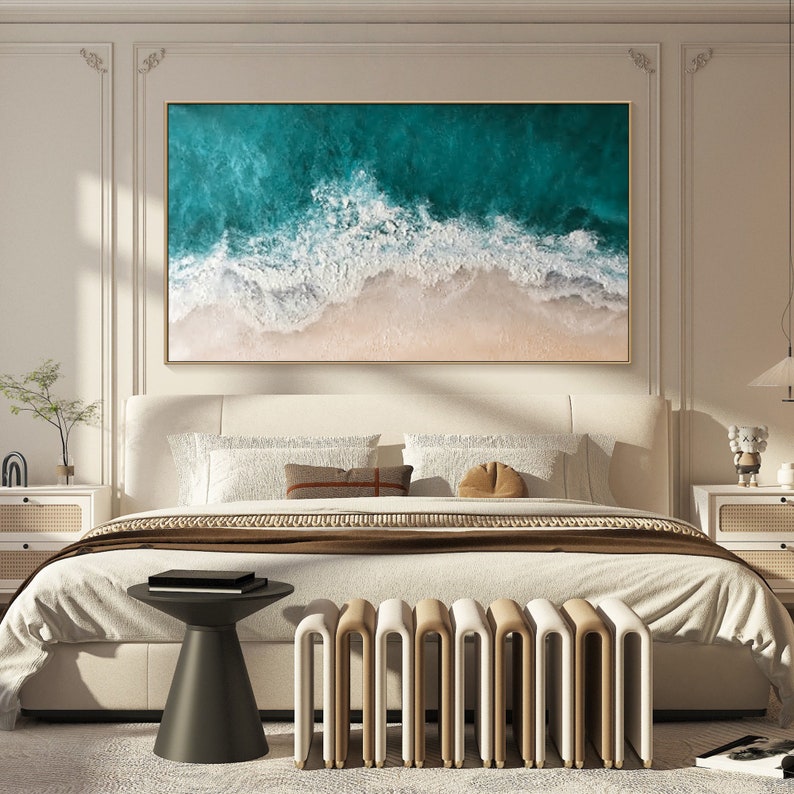 May include: A framed artwork depicting an aerial view of ocean waves crashing on a sandy beach. The painting features shades of teal and white for the water, and beige for the sand. The artwork is displayed above a bed in a bedroom setting.