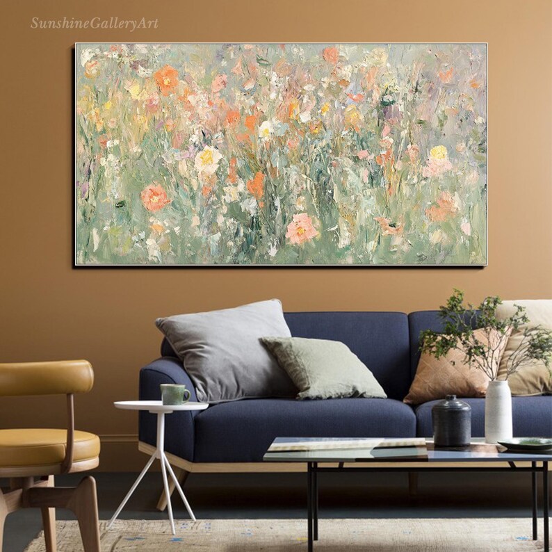 Op de afbeelding: Een abstract bloemenschilderij met zachte groen-, perzik- en wittinten. Het kunstwerk heeft een textuur, impressionistische stijl, met zichtbare penseelstreken die diepte en beweging cre&euml;ren. Het schilderij hangt boven een blauwe bank.