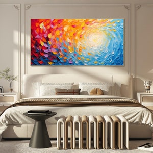 May include: Abstract painting with swirling colors of red, orange, yellow, blue, and purple. The painting is framed in gold and hangs above a white bed with a brown blanket and white pillows.