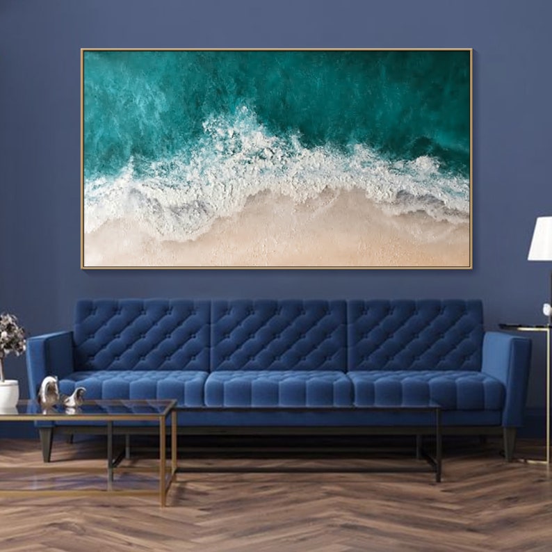 May include: Large framed artwork depicting an aerial view of a turquoise ocean meeting a sandy beach. The painting features textured white waves and a golden frame, creating a serene coastal scene. The artwork is displayed above a blue sofa.