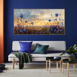 以下が含まれることがあります： 夕焼け空の下、青い花の咲く野原を描いた油絵。 金色のフレームに額装されたアートワークは、青と緑のクッションが付いた灰色のソファの上に飾られています。 緑色の毛布がソファに掛けられています。