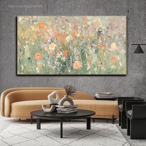 Op de afbeelding: Een abstract bloemenschilderij met een gouden lijst. Het kunstwerk toont een veld met bloemen in tinten oranje, roze, geel en wit tegen een groene en grijze achtergrond. Het schilderij wordt boven een gele bank getoond.