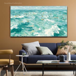 May include: An abstract painting of a turquoise ocean with numerous figures surfing and swimming. White waves and a light blue sky complete the scene. The artwork is framed and hangs above a blue sofa.