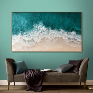 May include: A framed rectangular canvas art piece depicting an aerial view of a beach. The artwork features turquoise ocean waves crashing onto a sandy shore. The waves are textured with white foam, and the sand is beige. The frame is gold.