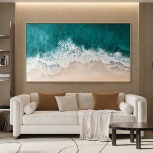 May include: Large framed artwork depicting an ocean scene. The painting features a teal and white ocean with foamy waves crashing onto a sandy beach. The artwork is displayed above a white sofa with brown and beige pillows.