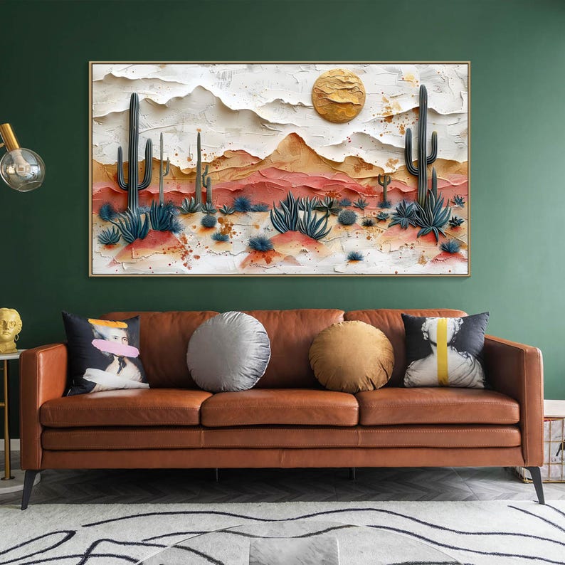 May include: A brown leather sofa with three cushions, two round and one rectangular. The sofa is in front of a green wall with a framed painting of a desert landscape with cacti and a golden sun.