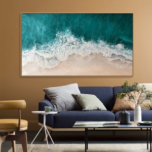 May include: A framed abstract painting depicting a beach scene. The artwork features a teal ocean with white foamy waves crashing onto a sandy shore. The painting is encased in a gold-colored frame, suitable for home decor.
