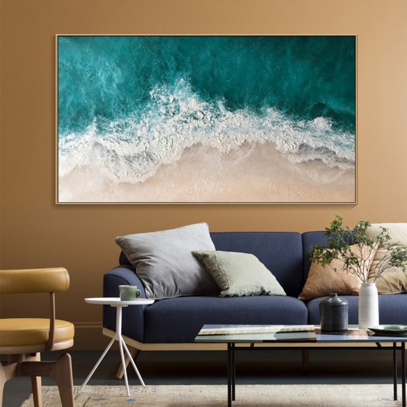 May include: A framed abstract painting depicting a beach scene. The artwork features a teal ocean with white foamy waves crashing onto a sandy shore. The painting is encased in a gold-colored frame, suitable for home decor.