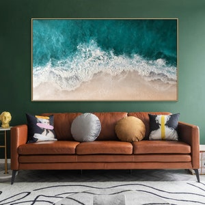 May include: Large framed artwork depicting an aerial view of ocean waves crashing on a sandy beach. The artwork is displayed above a brown leather sofa with decorative pillows. The room has a dark green wall and a patterned rug.