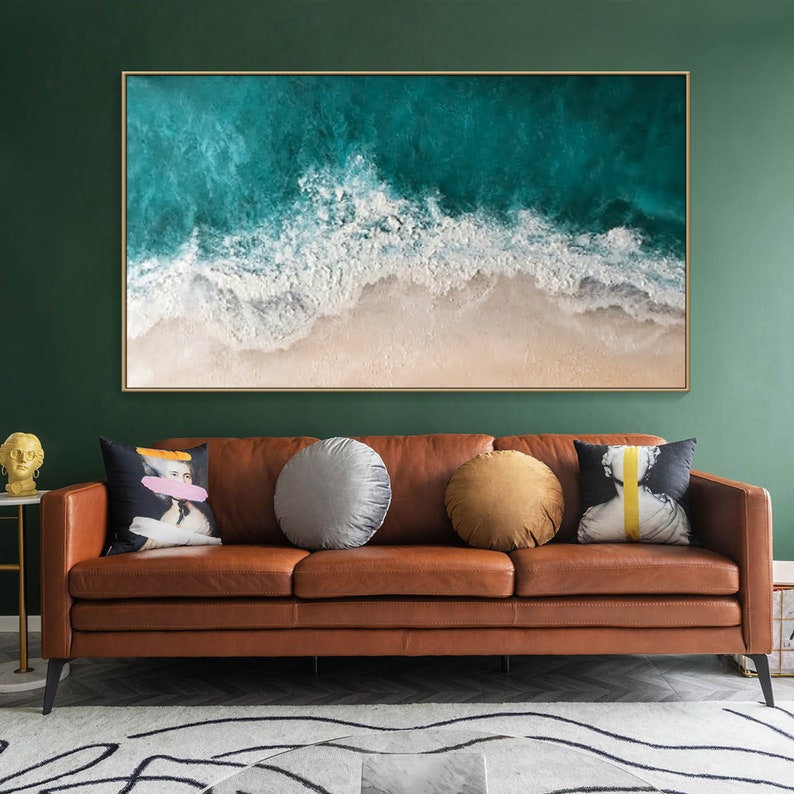 May include: Large framed artwork depicting an aerial view of ocean waves crashing on a sandy beach. The artwork is displayed above a brown leather sofa with decorative pillows. The room has a dark green wall and a patterned rug.