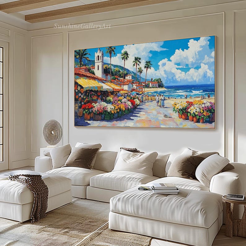 May include: A colorful painting of a seaside town with a white church, palm trees, and a flower market. The painting is framed and hanging on a white wall in a living room.