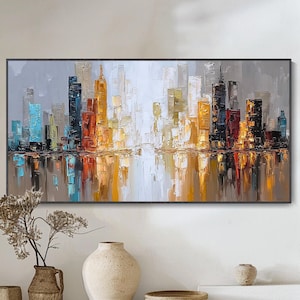 Puede incluir: Pintura enmarcada de un paisaje urbano con borde negro. La obra representa un horizonte con edificios coloridos en tonos de azul, naranja, amarillo y negro, reflejados en el agua de abajo. La pintura cuelga sobre un banco de madera con jarrones decorativos.