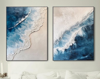 May include: Two abstract paintings of ocean waves crashing on a sandy beach. The paintings are in shades of blue, white, and beige. The paintings are framed in black frames.