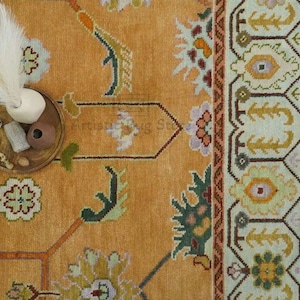 May include: A vintage rug with a light orange background and a geometric pattern in shades of green, brown, and white. The rug has a fringe border and is decorated with a small bowl and a white feather plume.
