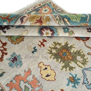 May include: A folded, light-colored rug with a detailed pattern. The rug features a cream-colored background with colorful floral and geometric designs in shades of teal, orange, yellow, and green. The edges of the rug are visible, showing the layered folds.