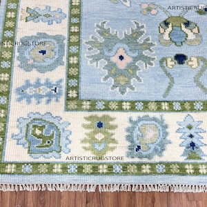 Op de afbeelding: Een lichtblauw tapijt met een witte achtergrond en groene, blauwe en roze bloemenpatronen. Het tapijt heeft een franje rand en de tekst "ARTISTICRUGSTORE" is drie keer op het tapijt gedrukt.