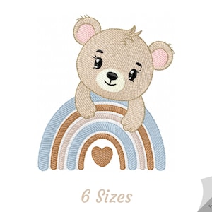 May include: Embroidered design of a brown bear peeking over a rainbow with a heart in the center. The rainbow is in shades of brown, blue, and white. The text "6 Sizes" is below the rainbow.
