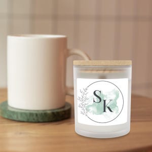Puede incluir: Una vela de vidrio esmerilado con tapa de madera, con una etiqueta blanca con las letras "SK" dentro de un c&iacute;rculo negro, un fondo aguamarina de acuarela y un dise&ntilde;o de hojas. Una taza blanca est&aacute; en el fondo.