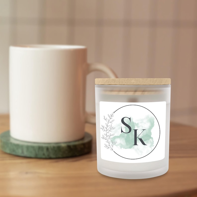 Puede incluir: Una vela de vidrio esmerilado con tapa de madera, con una etiqueta blanca con las letras "SK" dentro de un c&iacute;rculo negro, un fondo aguamarina de acuarela y un dise&ntilde;o de hojas. Una taza blanca est&aacute; en el fondo.