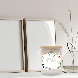 Puede incluir: Una vela perfumada blanca con tapa de madera, con un dise&ntilde;o floral y el texto "HELLO SUMMER". La vela est&aacute; rodeada por un marco decorativo y un jarr&oacute;n de cristal con vegetaci&oacute;n.