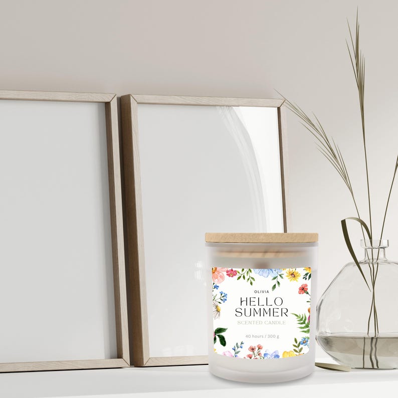 Puede incluir: Una vela perfumada blanca con tapa de madera, con un dise&ntilde;o floral y el texto "HELLO SUMMER". La vela est&aacute; rodeada por un marco decorativo y un jarr&oacute;n de cristal con vegetaci&oacute;n.