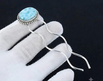 May include: A silver hair fork featuring a polished, oval-shaped light blue Larimar gemstone set in a beaded metal frame. The hair accessory has two long, wavy, silver-toned prongs that extend from the base of the stone setting.