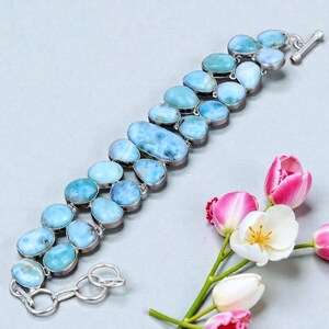 Pu&ograve; includere: Un bracciale a doppia fila con pietre ovali di larimar azzurro chiaro incastonate in argento. Il bracciale ha una chiusura a levetta ed &egrave; esposto con tulipani rosa e bianchi.