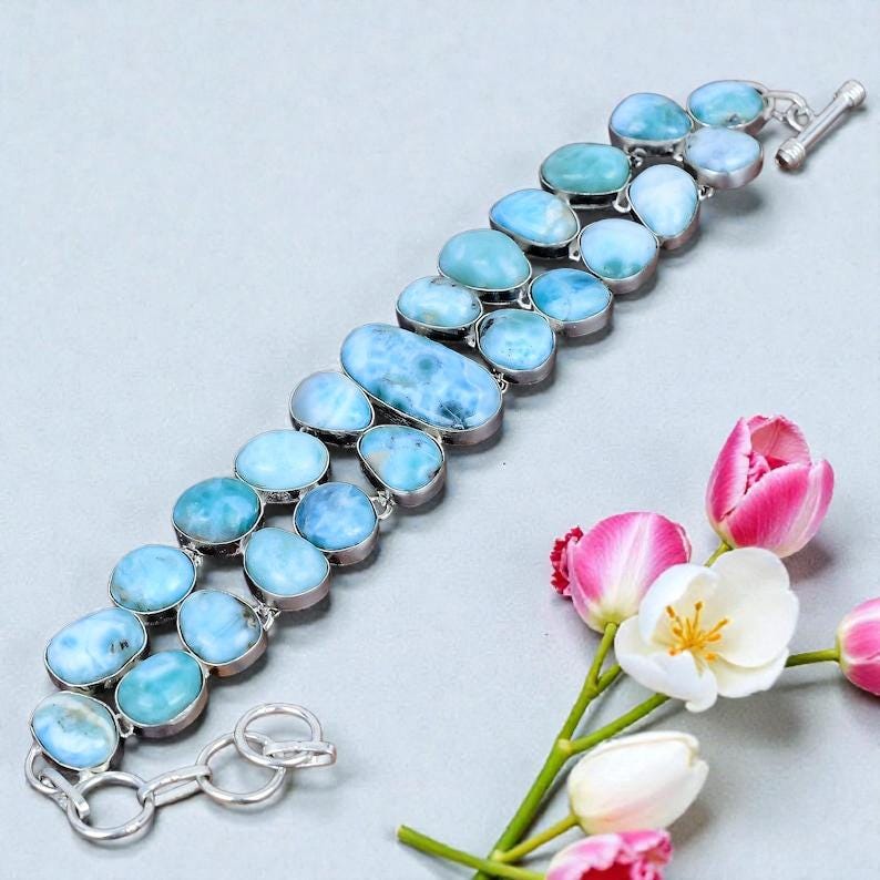 Pu&ograve; includere: Un bracciale a doppia fila con pietre ovali di larimar azzurro chiaro incastonate in argento. Il bracciale ha una chiusura a levetta ed &egrave; esposto con tulipani rosa e bianchi.