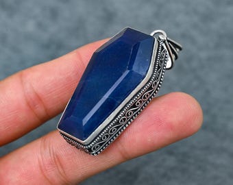 May include: A dark blue coffin-shaped pendant with a silver filigree border. The pendant is a deep, faceted blue, and the silver setting has an ornate, swirling design. The bail is a simple loop for a chain. The pendant is held in a hand.