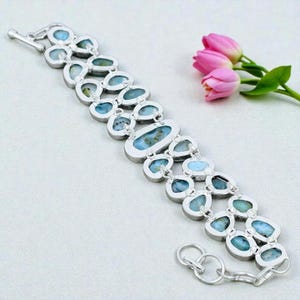 Pu&ograve; includere: Un bracciale in argento con pi&ugrave; pietre preziose blu ovali e a goccia. Il bracciale ha un design a doppia fila con una chiusura sicura. Tulipani rosa sullo sfondo.