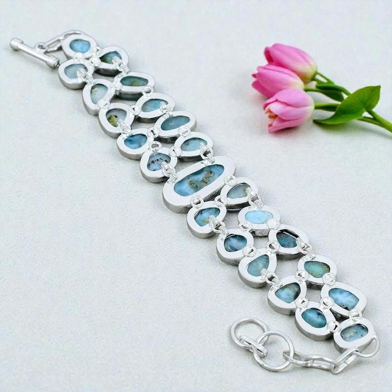 Pu&ograve; includere: Un bracciale in argento con pi&ugrave; pietre preziose blu ovali e a goccia. Il bracciale ha un design a doppia fila con una chiusura sicura. Tulipani rosa sullo sfondo.