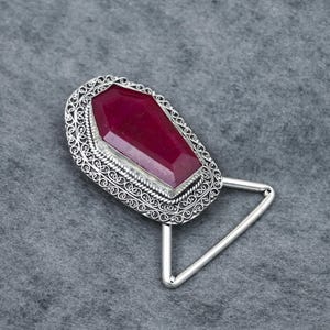 May include: A silver-toned bookmark with a large, rectangular, faceted, deep red gemstone. The gemstone is set in a decorative, filigree-style frame. The bookmark has a triangular base.