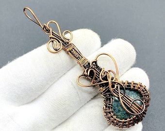 May include: A copper wire wrapped pendant featuring a green stone in the shape of a guitar. The pendant has a loop at the top for attaching to a chain.