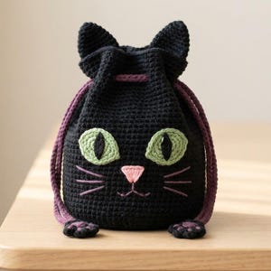 May include: A black crochet drawstring pouch shaped like a cat. The bag features two pointed ears, large green eyes with black pupils, a small pink embroidered nose, and purple whiskers. Two purple braided drawstrings cinch the top of the bag.