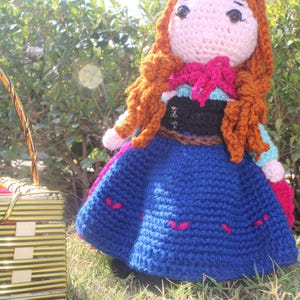 K&ouml;nnte beinhalten: Eine geh&auml;kelte Puppe mit langen braunen Haaren, die ein blaues Kleid mit rosa Akzenten und einen braunen G&uuml;rtel tr&auml;gt. Die Puppe steht auf gr&uuml;nem Gras.